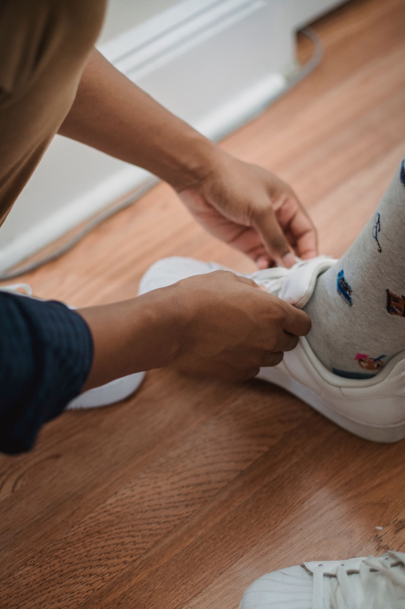 A parent tying a child's white sneaker on a wood floor.